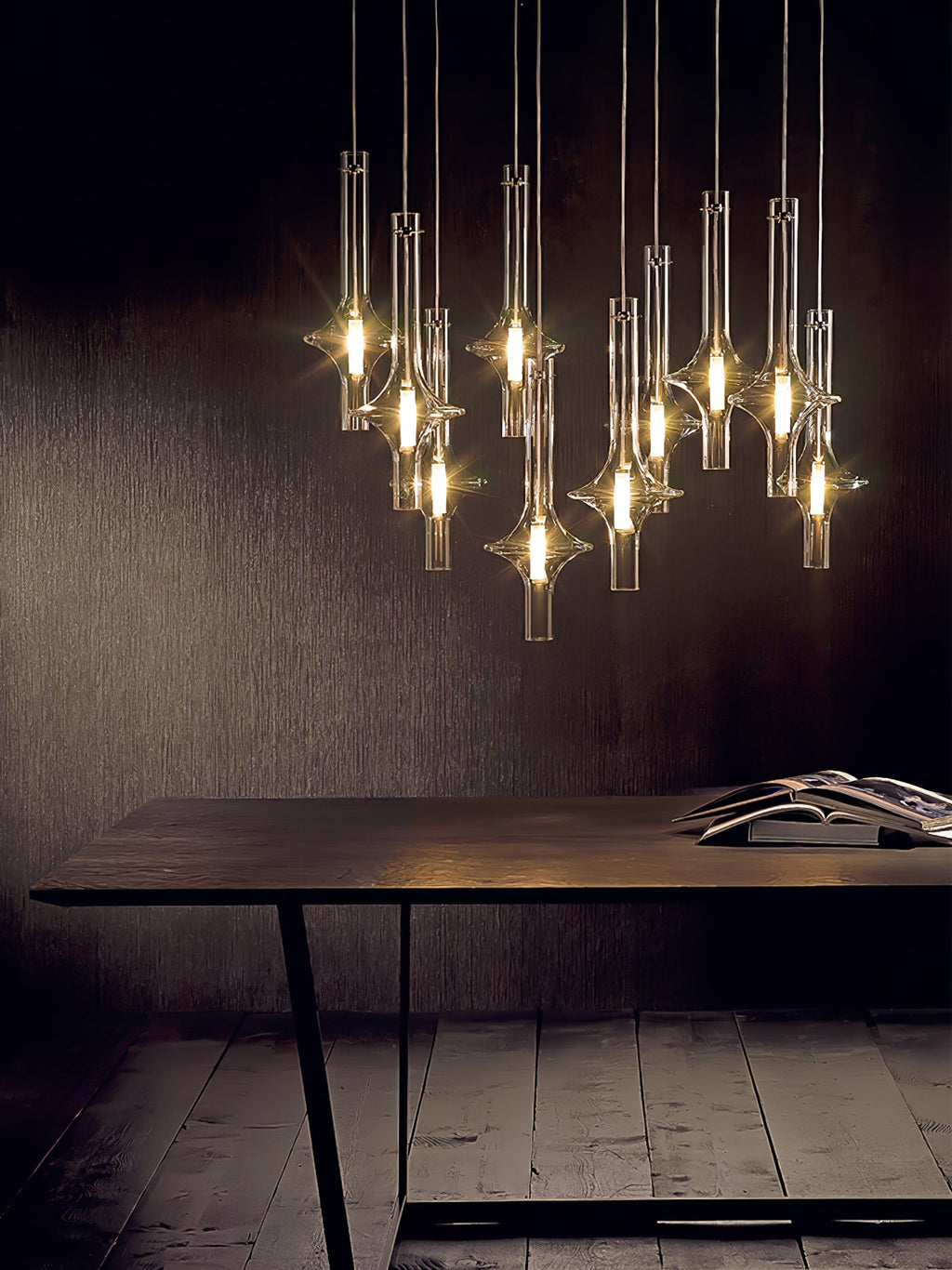 A cluster of lit Clérachic pendants hangs above a dark wood table with an open book, against a dark studio wall.