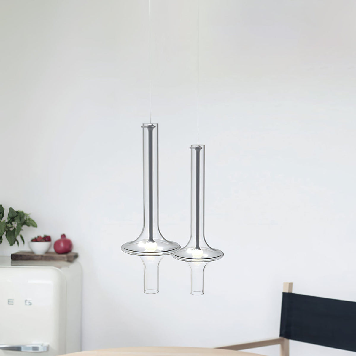 Two clear-glass Clérachic pendants hang in a white kitchen beside a cream retro fridge and a black chair.