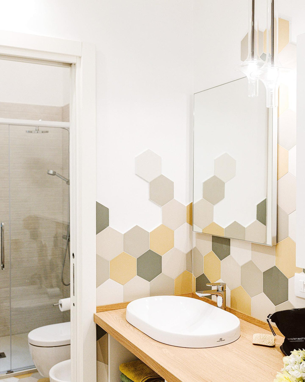 A single lit Clérachic pendant hangs in a bathroom lined with hexagon-patterned tiles, beside a mirror and a white basin.