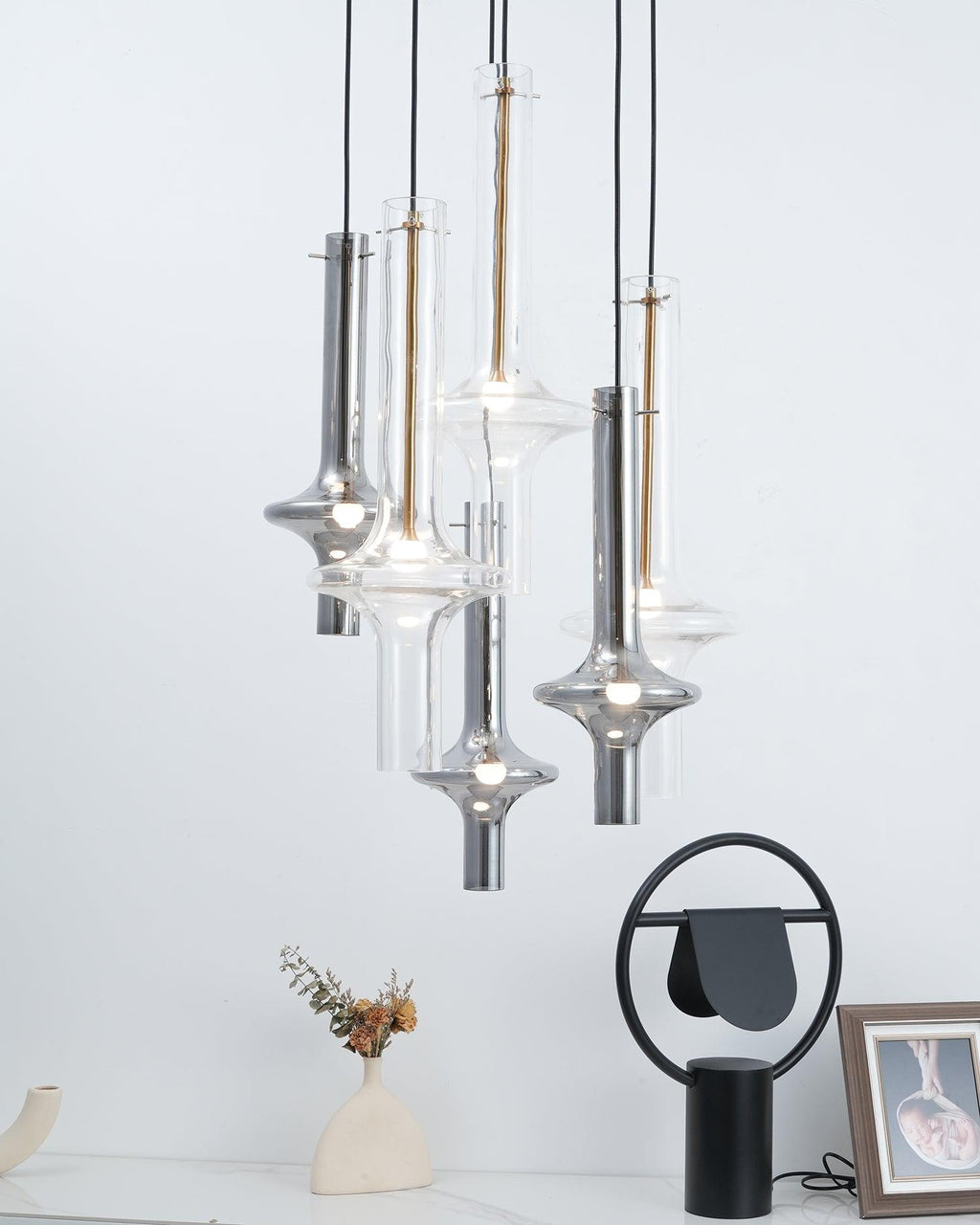 A cluster of gold, black and clear-stemmed Clérachic pendants hangs above a round side table with a vase and an orange strap chair.