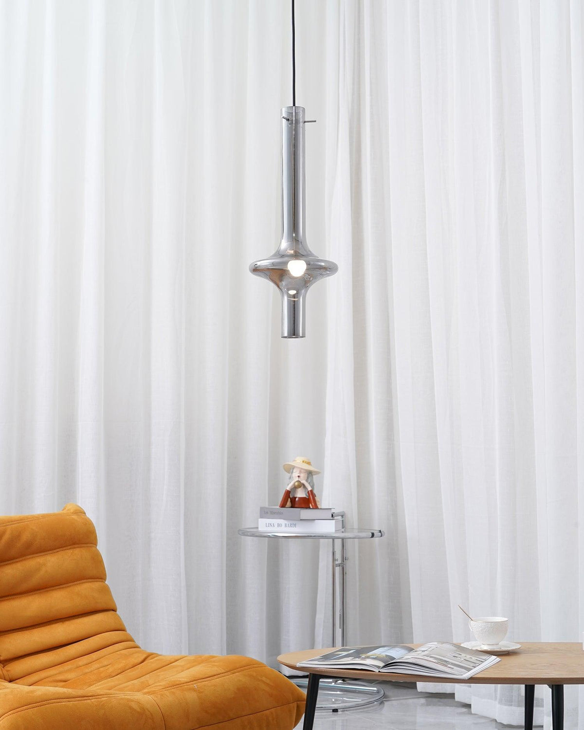 A single dark-stemmed Clérachic pendant glows against sheer white curtains, above a round side table beside a mustard armchair.