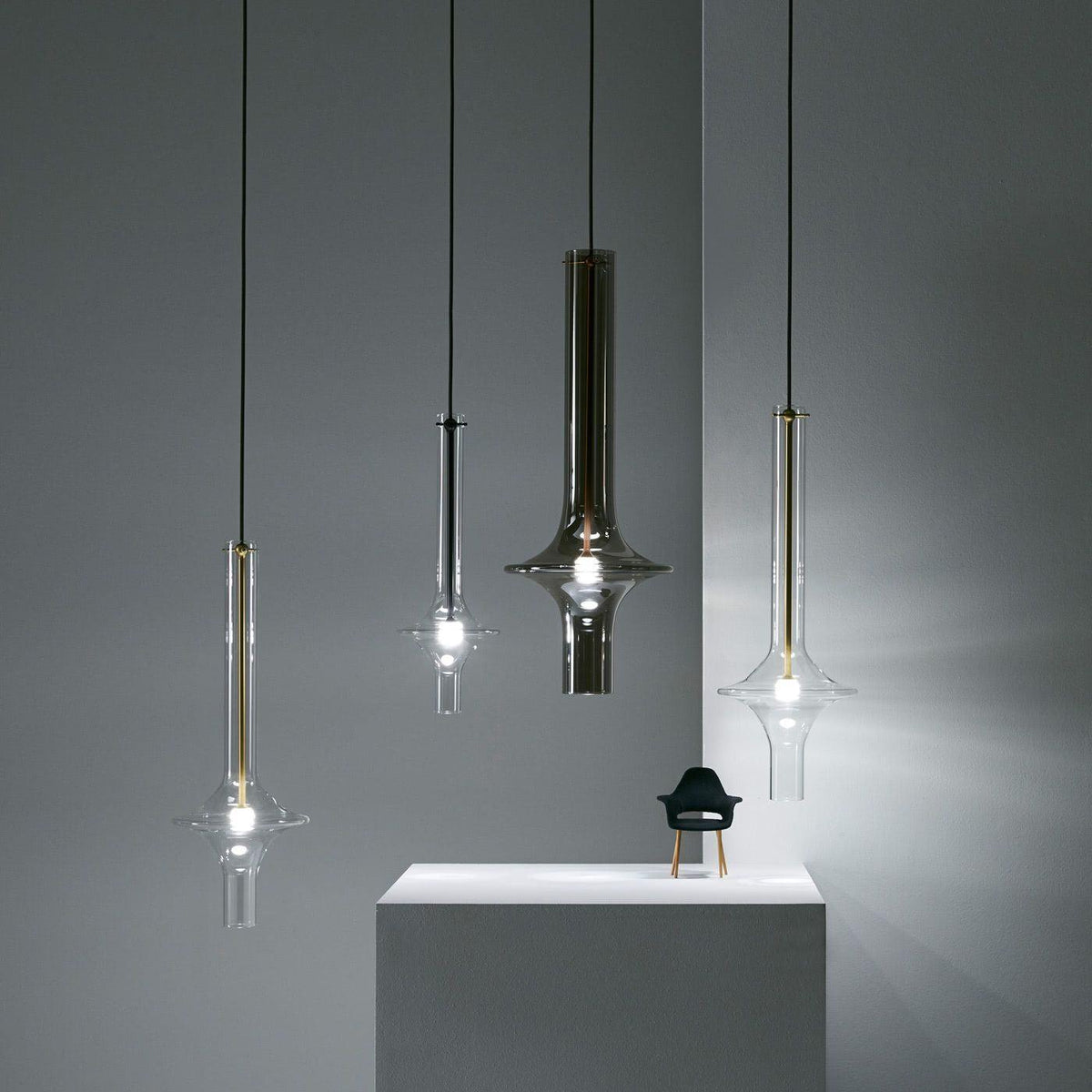 Four single Clérachic pendants in black, smoky and clear glass hang at staggered heights beside a small chair figurine on a plinth.