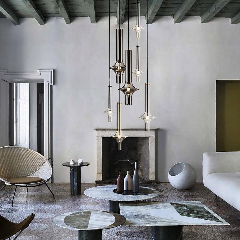 A cluster of lit Clérachic pendants hangs over a stone fireplace mantel in a rustic room with a woven chair and round marble tables.