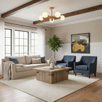 Beneath exposed wood ceiling beams in a living room with a cream sofa and blue armchairs, this Clérachic chandelier glows warmly.