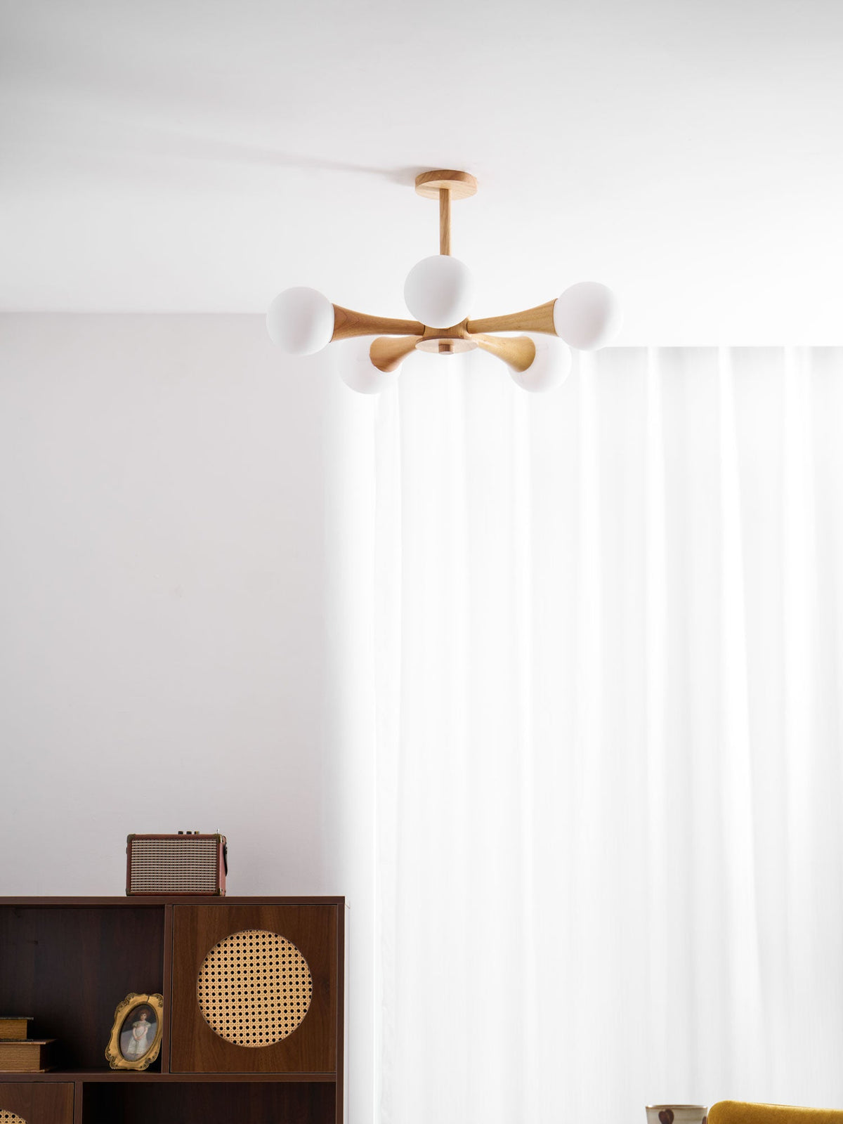 In a softly lit corner with sheer curtains, this Clérachic chandelier's pale wood arms and opal glass globes glow above a dark wood cabinet.
