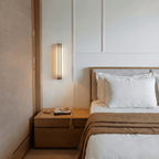 Clérachic marble and copper wall light glowing above an oak bedside table set with a strand of wooden beads, on a white panelled wall.