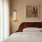 Clérachic marble and copper wall light lit beside a curved dark-wood headboard, between a sheer curtain and a framed botanical print.