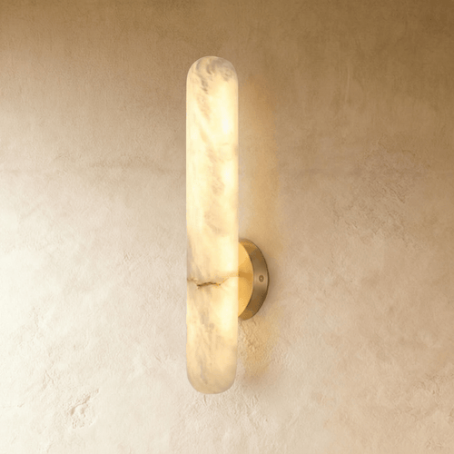 Clérachic alabaster wall lamp casting a warm amber glow across a rough beige plaster wall, its light spilling from a slender vertical stone bar.