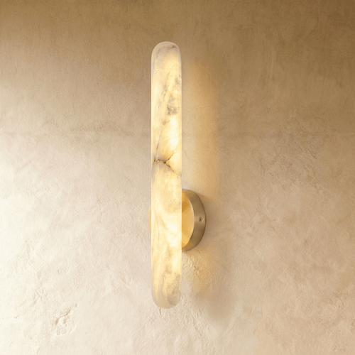 Clérachic alabaster wall lamp lit on a warm plaster wall, its grey striations showing through the glowing stone and a halo of light spreading behind it.