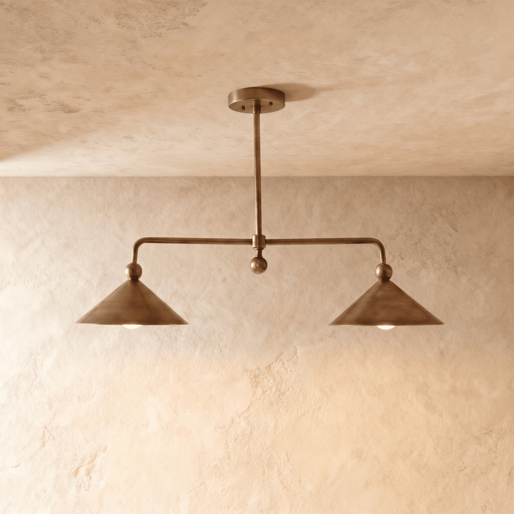 Clérachic chandelier in vintage brass, two angled cone shades hung from a slim rod against a plaster wall.