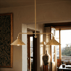 Clérachic chandelier in brass hanging over a sunlit living room with a tapestry, doorway and vase.