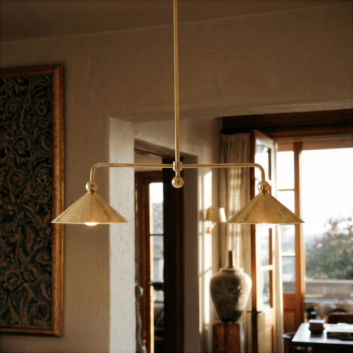 Clérachic chandelier in brass hanging over a sunlit living room with a tapestry, doorway and vase.