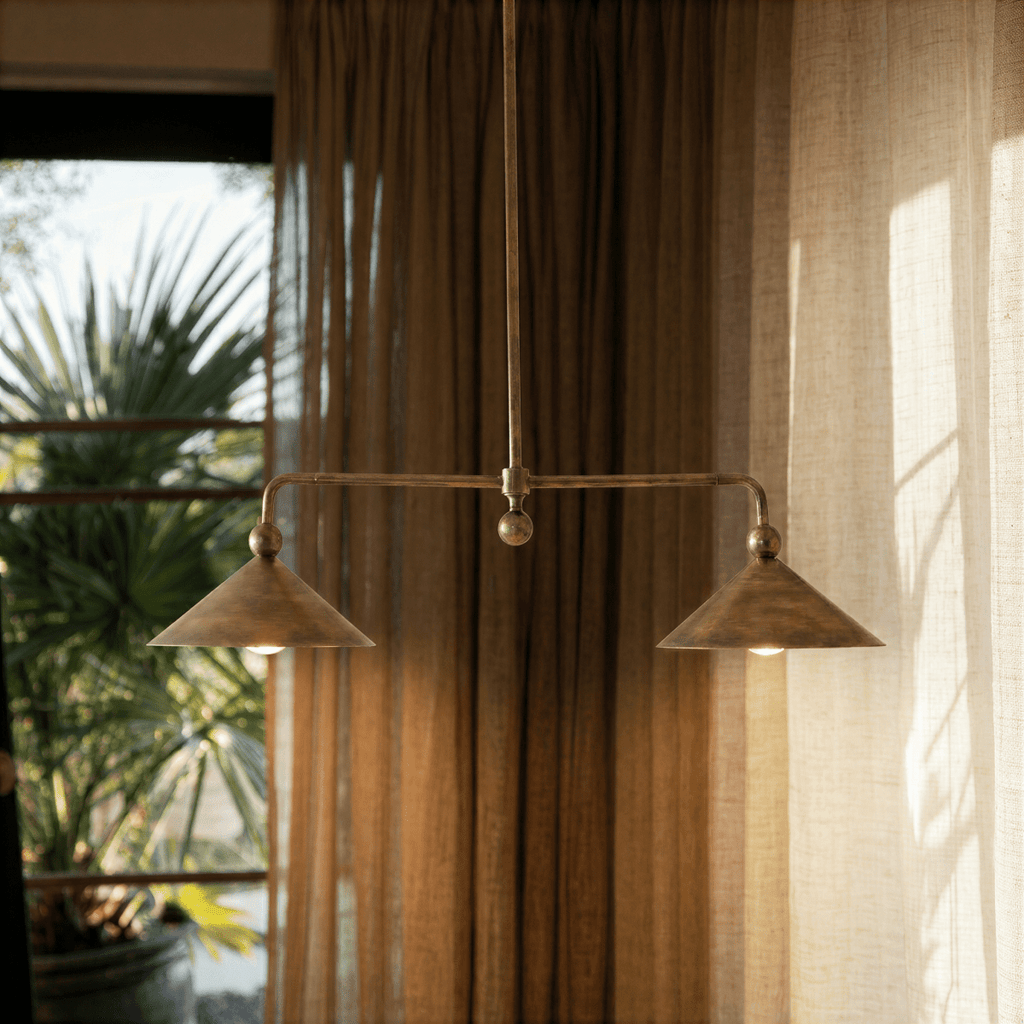 Clérachic chandelier in brass hanging in front of soft curtains beside a sunlit palm-view window.
