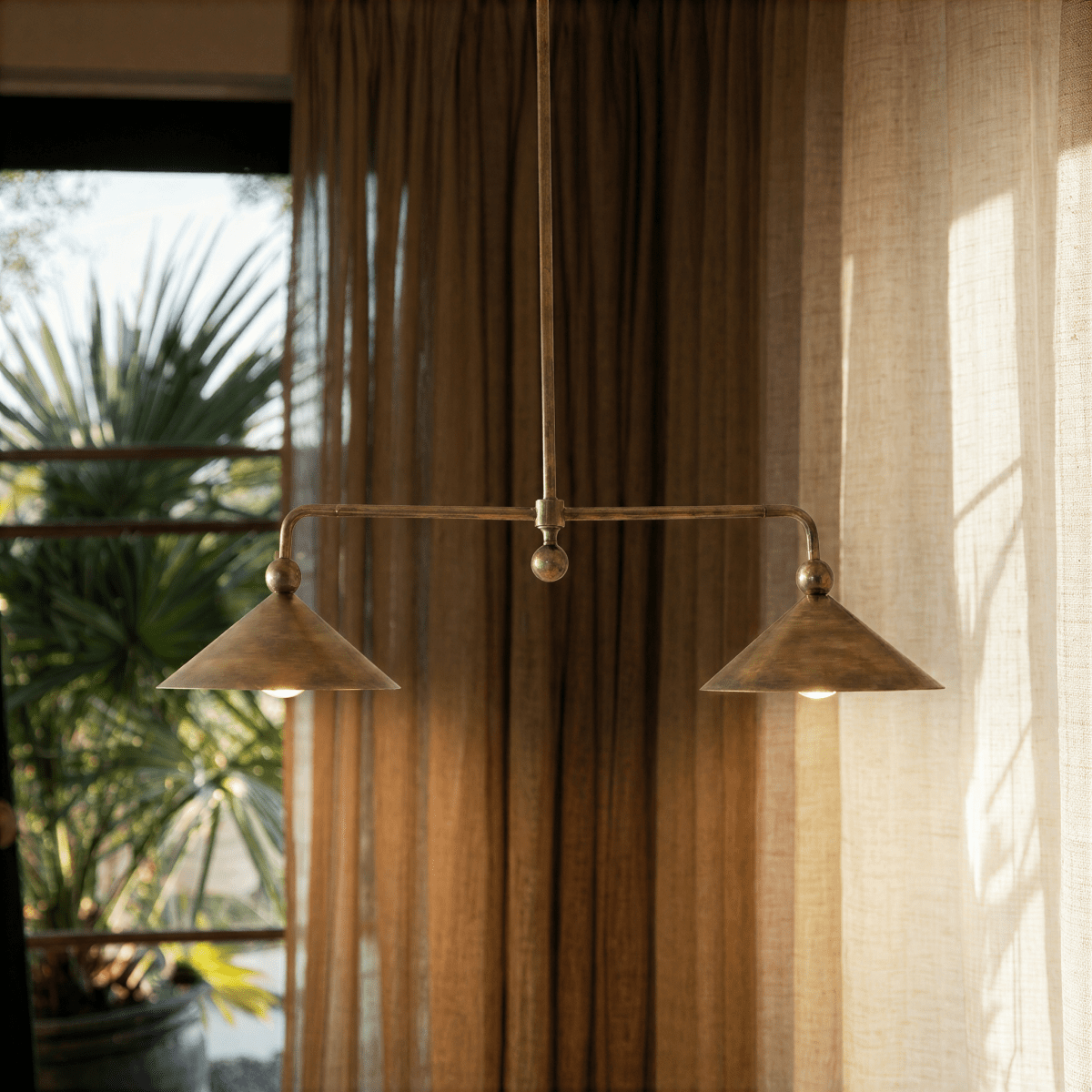 Clérachic chandelier in brass hanging in front of soft curtains beside a sunlit palm-view window.