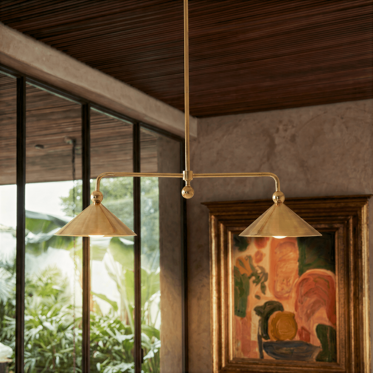 Clérachic chandelier in brass hanging beside a framed painting and a window looking onto greenery.