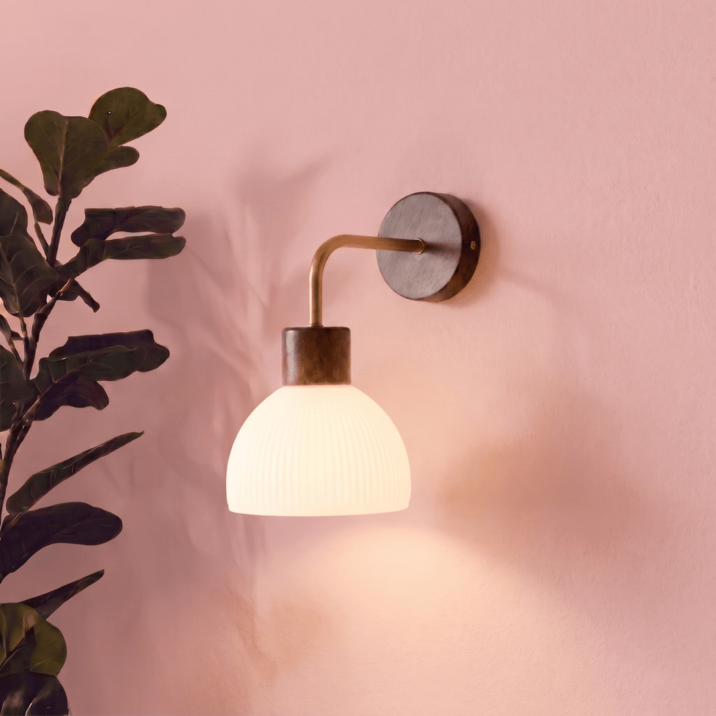 This Clérachic wall light glows softly on a pink wall beside a leafy potted plant, its glass dome lit warm.