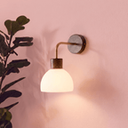 This Clérachic wall light glows softly on a pink wall beside a leafy potted plant, its glass dome lit warm.