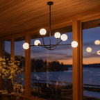Clérachic six-globe chandelier in a matte black finish, its globes glowing warm white at dusk in the same wood-lined room, with an orange sky over the water beyond the windows.