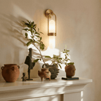 Clérachic brass wall lamp lit above a pale mantel styled with terracotta pots, dark figurines and a leafy branch.