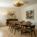 Clérachic brass flush ceiling light glowing above an oval dining table beneath two large colourful abstract paintings.