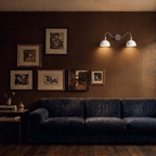 Clérachic twin wall light in chrome, mounted on a deep brown wall beside a gallery of framed prints, lighting the end of a blue velvet sofa in a dim living room.