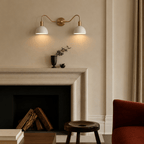 Clérachic twin wall light on a plaster wall above a stone fireplace mantel, lighting a dark footed urn holding a bare branch and a small dark bowl beside it.
