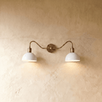 Clérachic twin wall light in vintage brass, lit, its darker antiqued rod above two white domes that drop a soft pool of light onto the plaster wall.