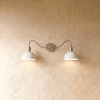 Clérachic twin wall light in chrome, lit, the cool silver rod and collars set against warm plaster between two pools of light.