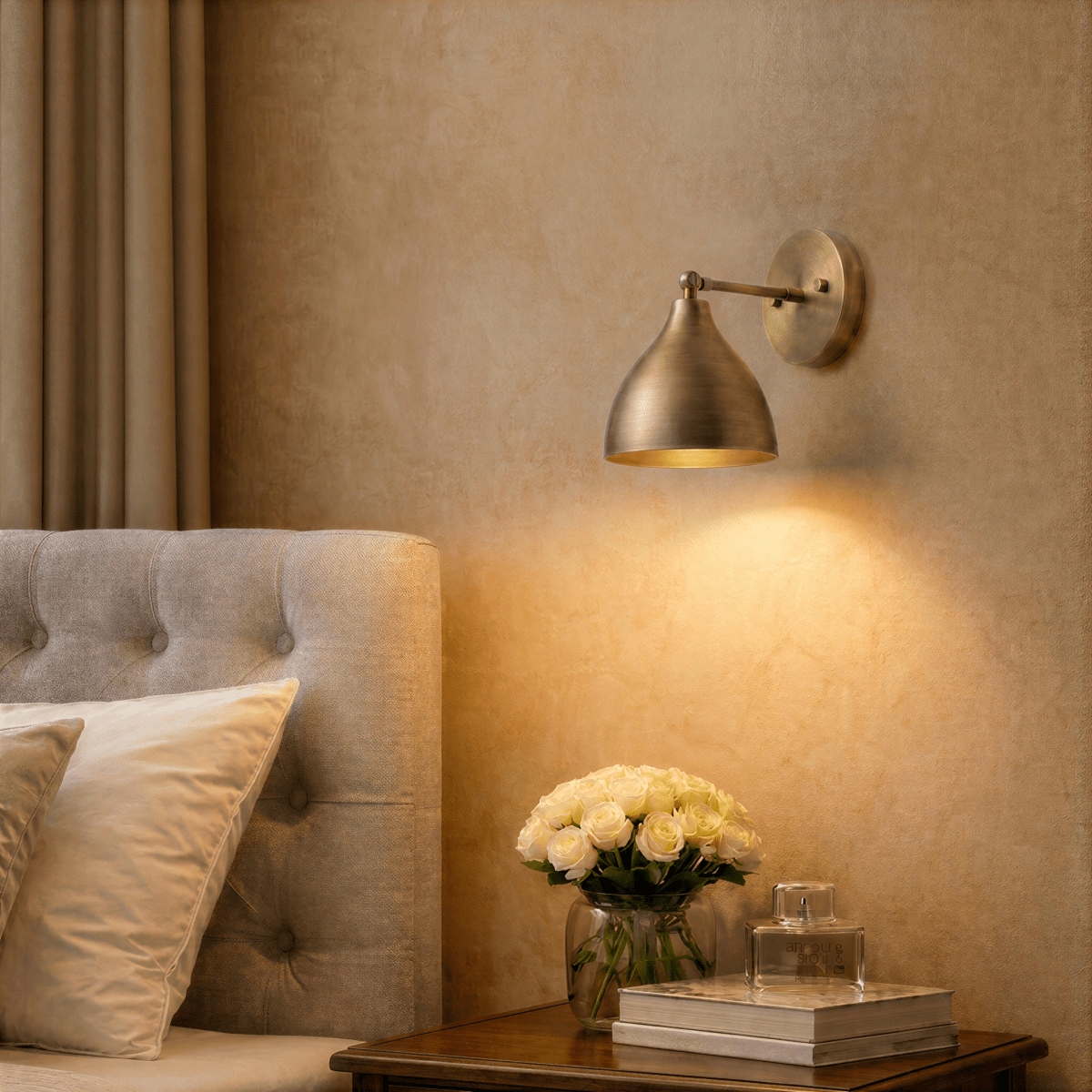 A vintage brass Clérachic wall lamp lights a bedroom nightstand holding roses and a glass perfume bottle.