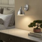 A chrome Clérachic wall lamp lights a marble-top nightstand holding a bonsai and stacked books.