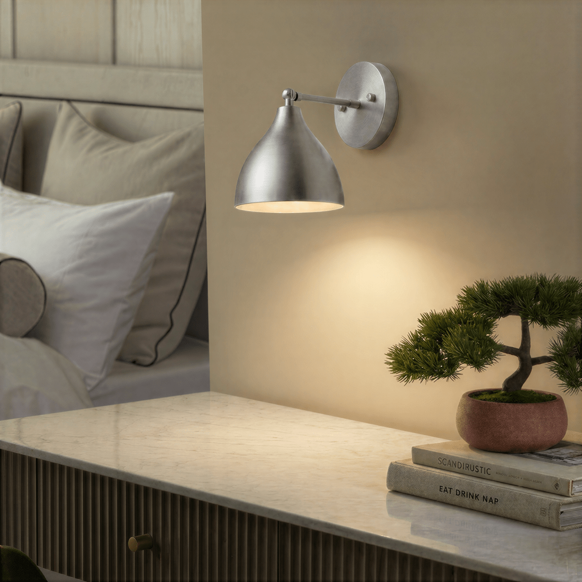 A chrome Clérachic wall lamp lights a marble-top nightstand holding a bonsai and stacked books.