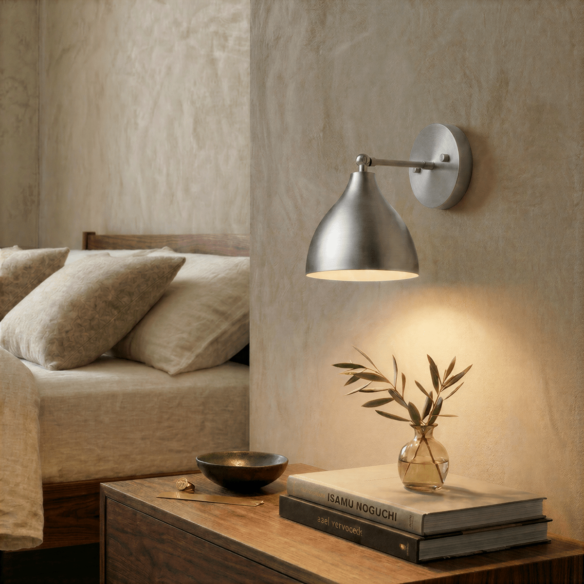 This chrome Clérachic wall lamp lights a bedroom nightstand stacked with books beside a small glass vase.