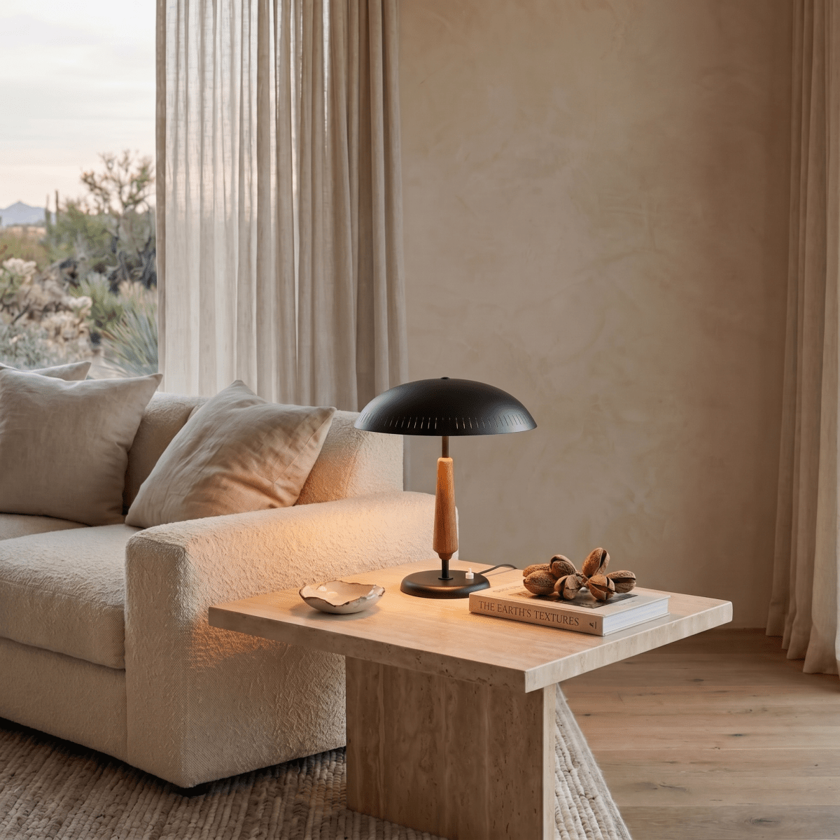 Clérachic matte black table lamp glowing on a travertine side table beside a beige sofa and desert-view window.