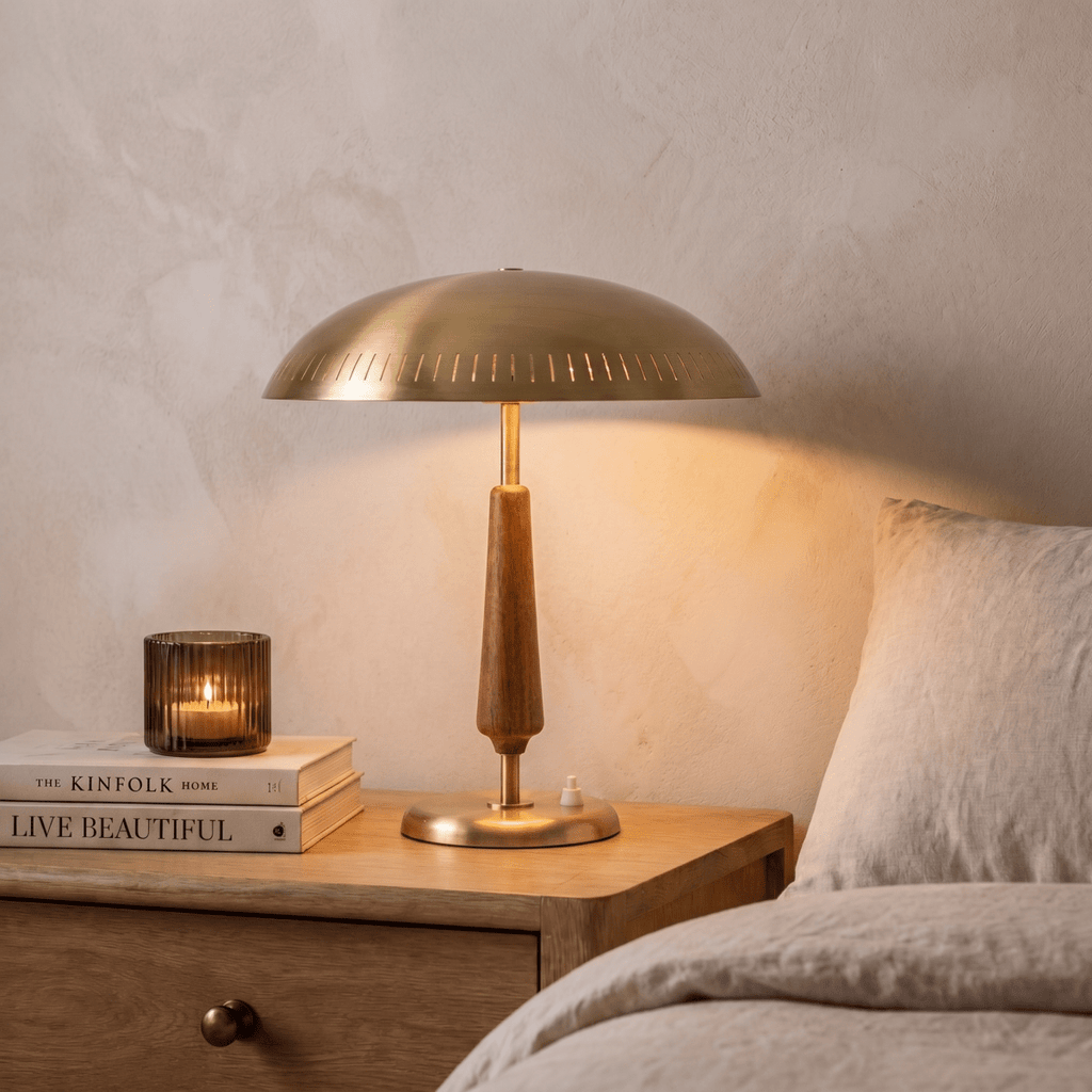 Clérachic brass table lamp glowing on a bedside table stacked with books, beside a linen-dressed bed.
