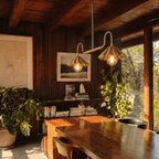 Clérachic double-arm ceiling chandelier in brass, hung above a long wooden table between potted plants and a shelf of stacked books.