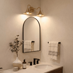 Clérachic brass double-head wall sconce mounted above an arched mirror beside a hanging linen towel in a bathroom.