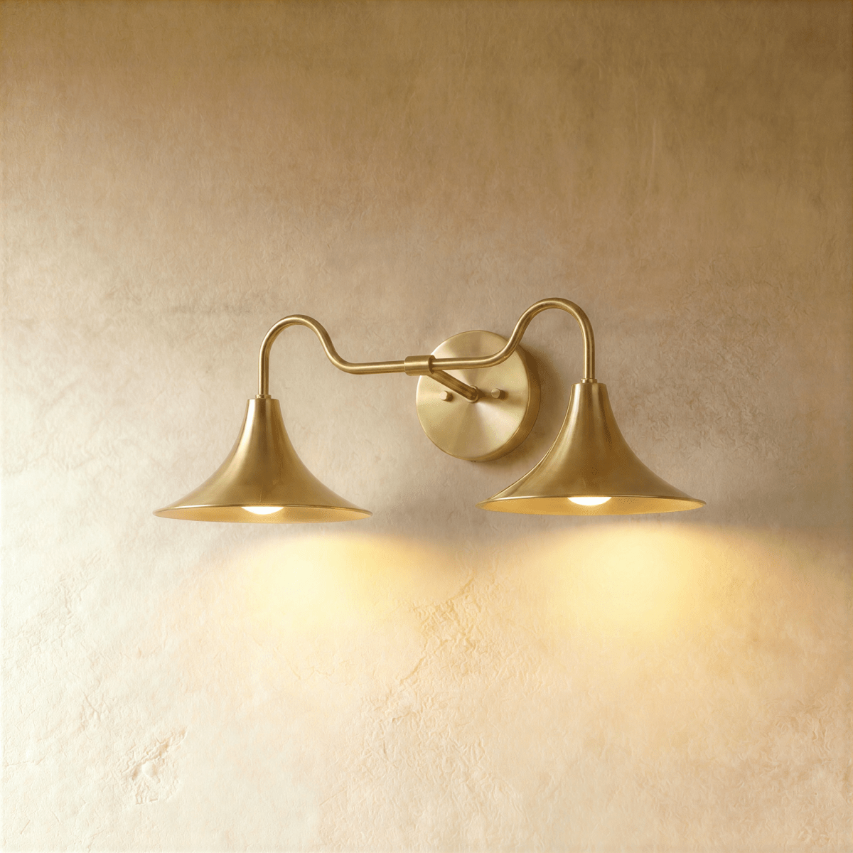 Clérachic double-head wall sconce in a brighter brass finish, its trumpet shades curving outward on a plaster wall.