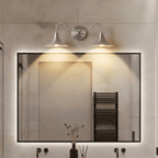 Clérachic chrome double-head wall sconce glowing above a backlit mirror in a tiled bathroom with a towel warmer.