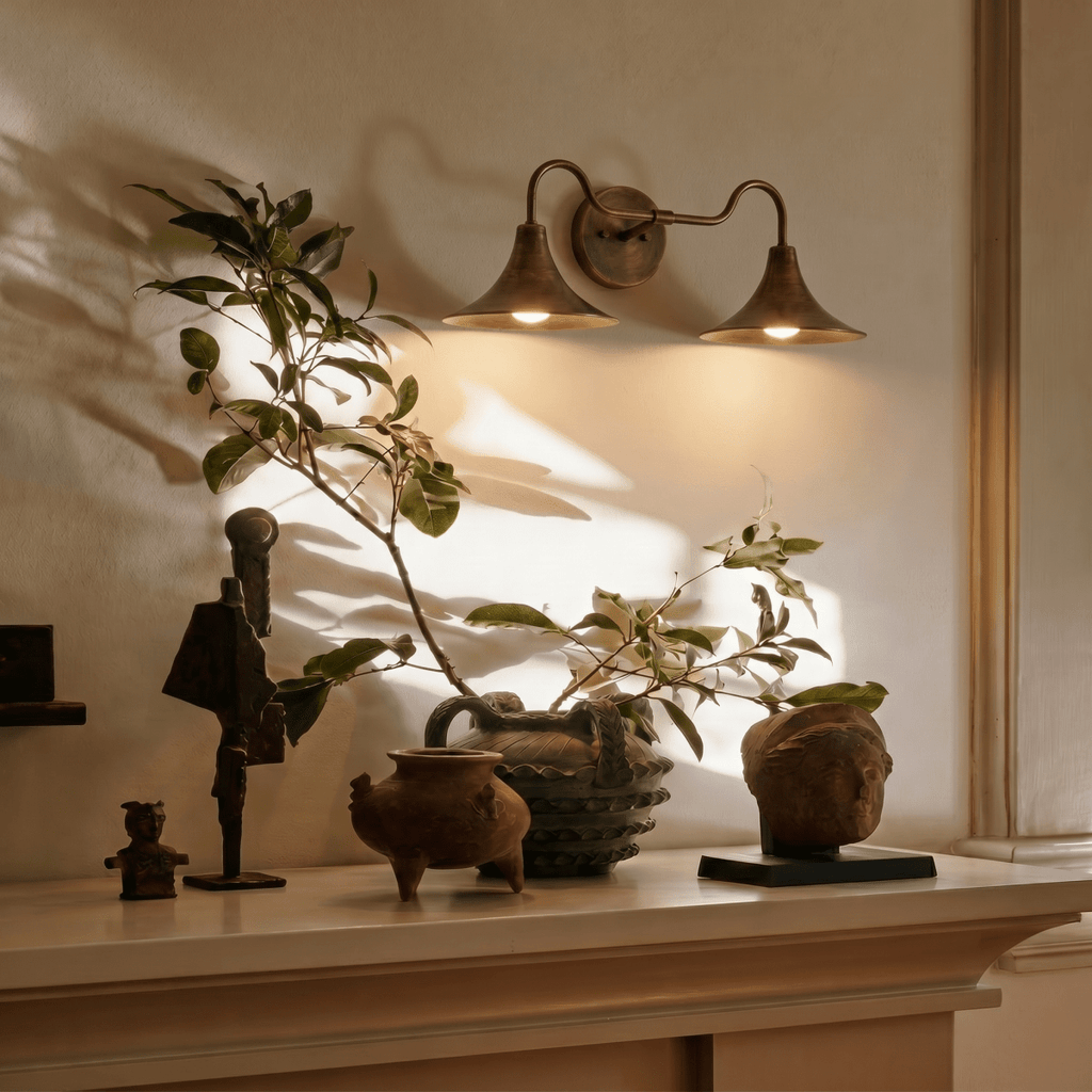 Clérachic vintage brass double-head wall sconce glowing above a mantel styled with carved figures and potted greenery.