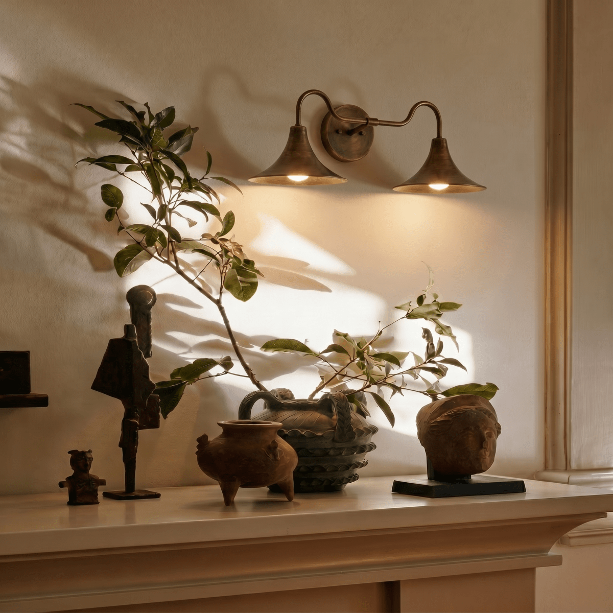 Clérachic vintage brass double-head wall sconce glowing above a mantel styled with carved figures and potted greenery.