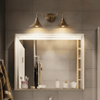Clérachic brass double-head wall sconce glowing above a framed mirror beside a towel and folded bathrobe.