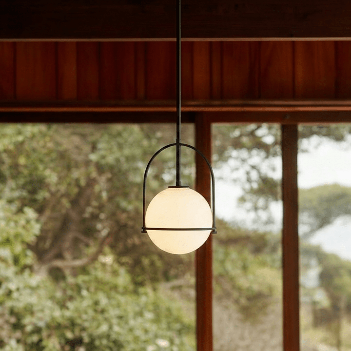 Clérachic globe pendant light in matte black, lit against dark wood panelling with greenery visible through the glass behind it.