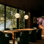 Clérachic globe pendant lights in matte black, paired above a dining table with patterned velvet chairs and a lake framed by the glass wall.