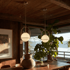 Clérachic globe pendant lights in brass-tone gold, paired above a wood table set with a weathered clay vessel and a large-leafed plant, water beyond.