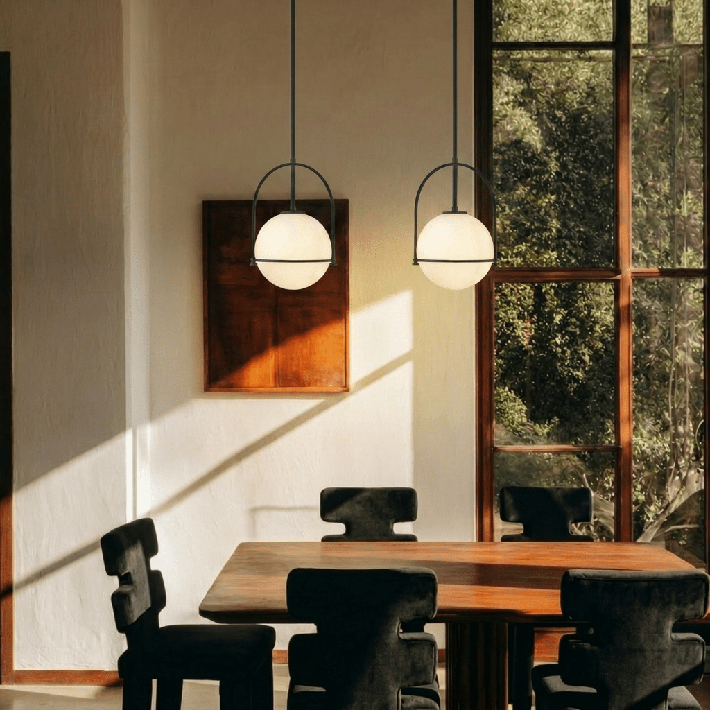 Clérachic globe pendant lights in matte black, paired above a dining table with dark sculptural chairs, sunlight raking across the plaster wall behind.