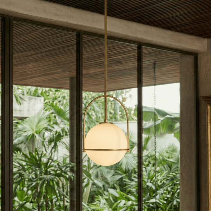 Clérachic globe pendant light in brass-tone gold, glowing close to floor-to-ceiling windows framed by tropical foliage.