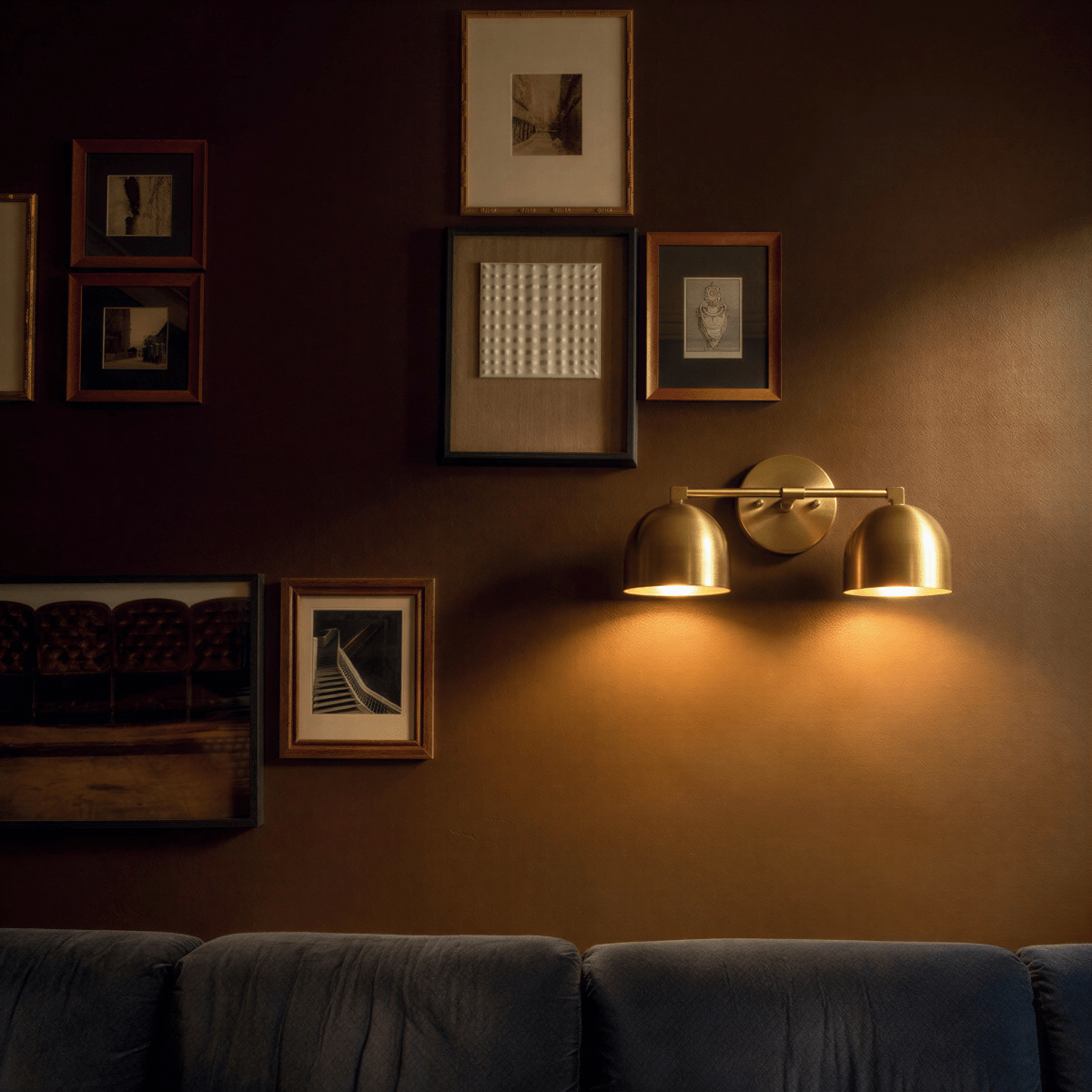Clérachic double wall lamp in brass, lit on a dark wall between a gallery hang of framed prints and the sofa below.