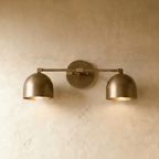 Clérachic double wall lamp in vintage brass, its darker, greyer tone shown lit against a plaster wall.
