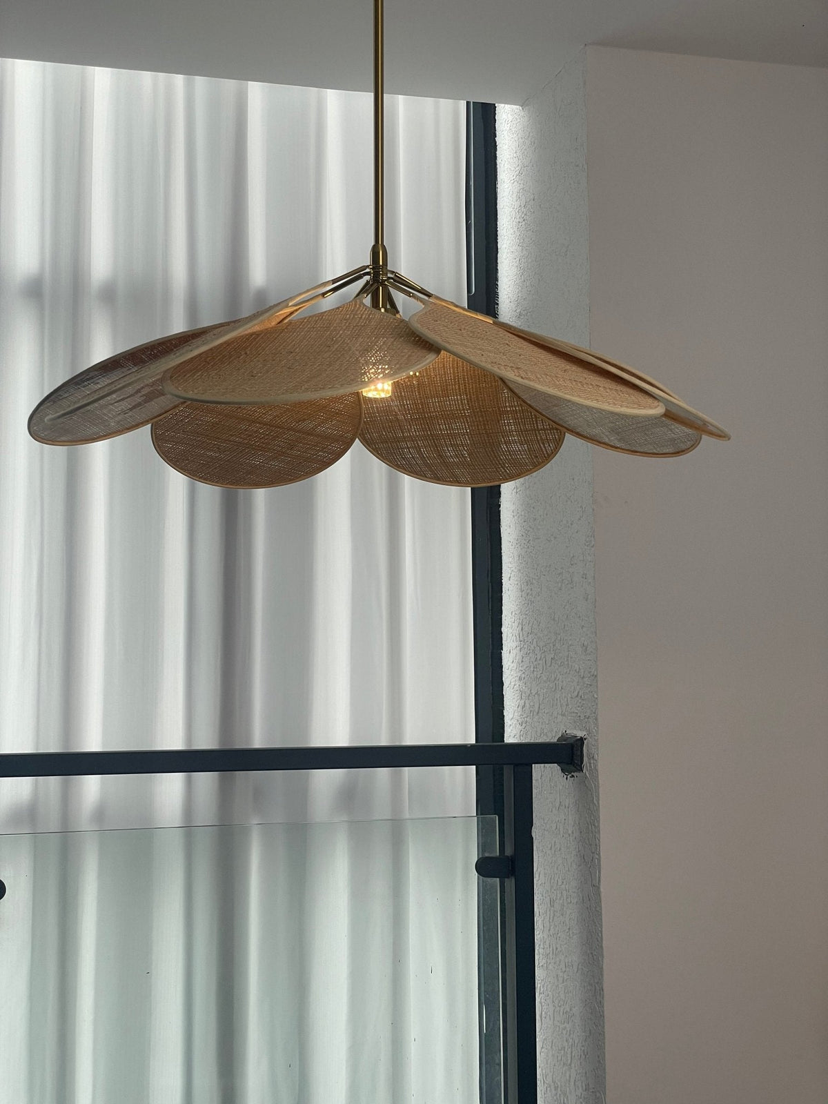 Close view of this Clérachic pendant lamp's natural woven rattan petals, hanging beside a curtained window.