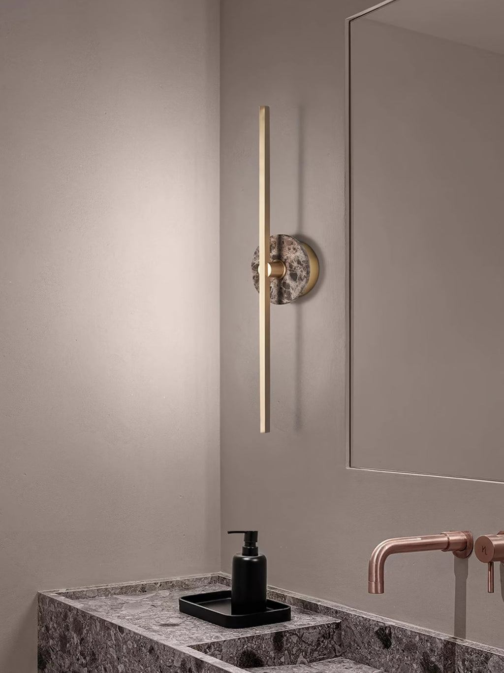 Clérachic wall light mounted beside a bathroom mirror, its brass bar rising above a marble vanity with copper taps.
